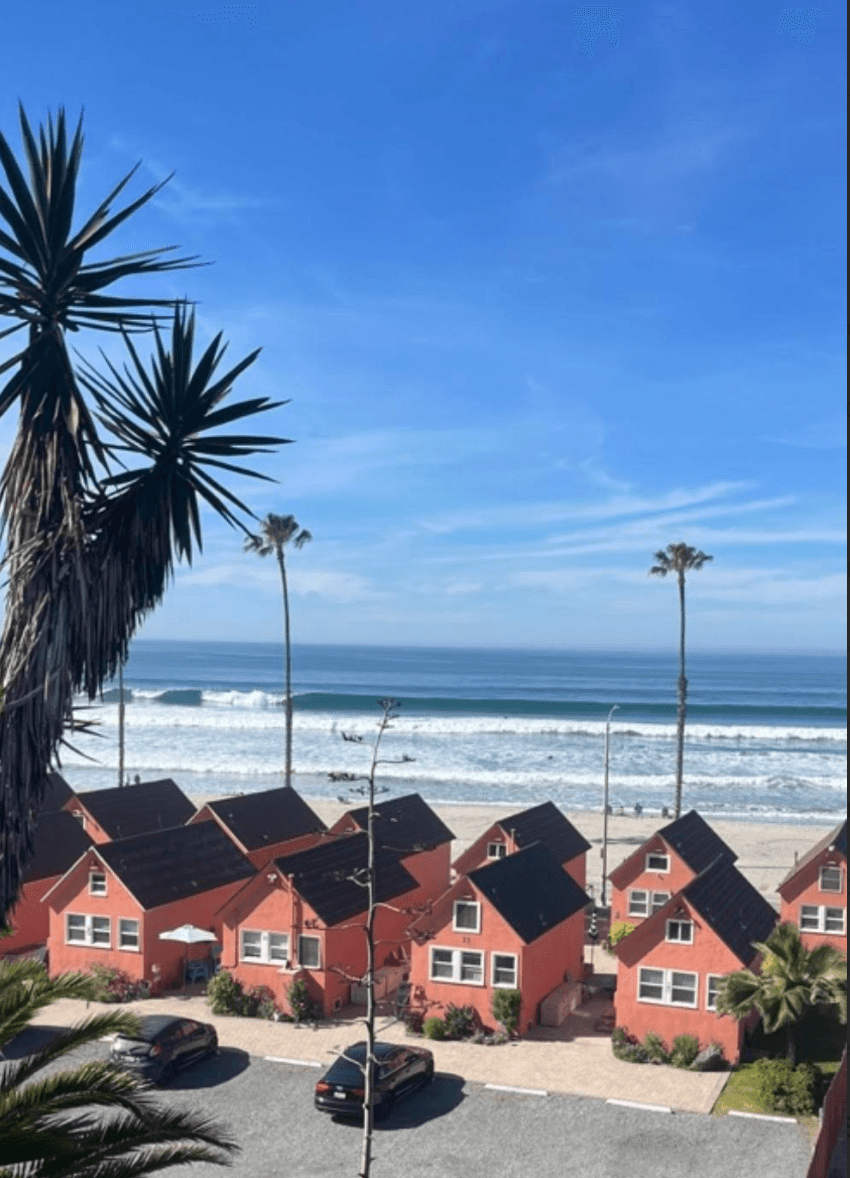Small pink cottages overlook a sandy beach with rolling ocean waves and palm trees.