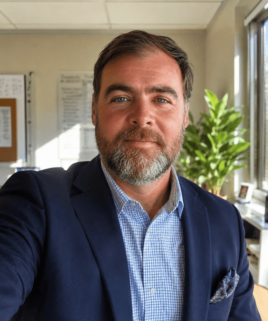 Bearded man in a navy blazer and checkered shirt, smiling in a bright office.